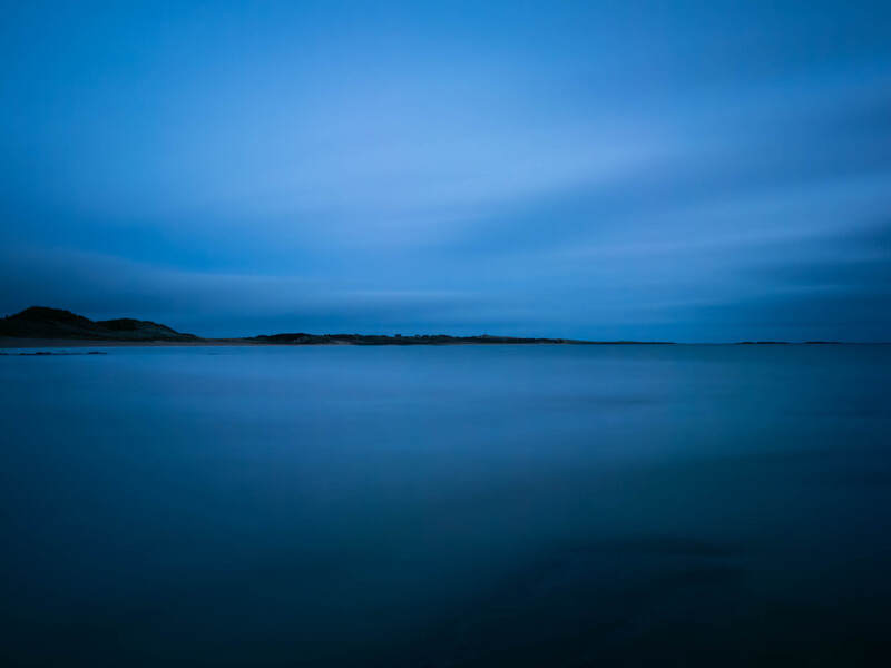 Serene seascape at twilight with calm, mirror-like water under a deep blue sky.