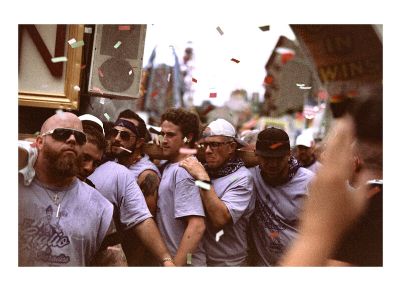 Group of men wearing white t-shirts and caps gathered on an urban street, with confetti falling around them.