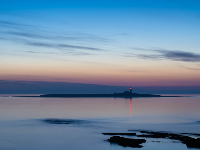 Silhouetted lighthouse on a distant peninsula at twilight with calm, mirrored water and rocky foreground.