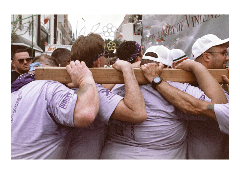 Group of people in lavender shirts carrying a wooden beam across their shoulders at an outdoor event.