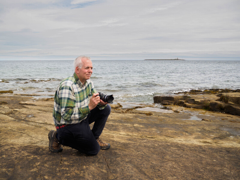 Photographer crouching on rocky beach while holding a camera, facing the ocean under cloudy sky.