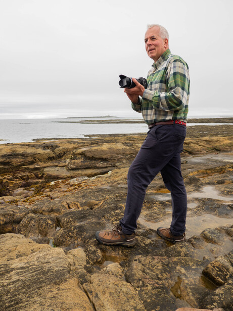 Photographer holding a mirrorless camera while standing on rocky coastal terrain with calm water and overcast sky behind him.