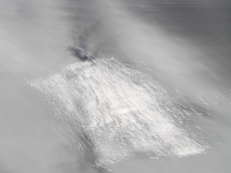 Long exposure photograph of a person in motion with blurred white fabric against a soft gray background.