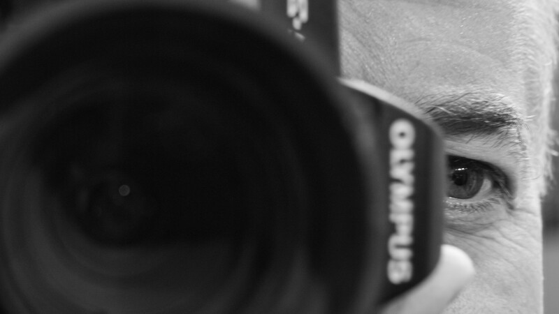 Photographer holding a camera with telephoto lens up to their face, revealing one eye in black and white.