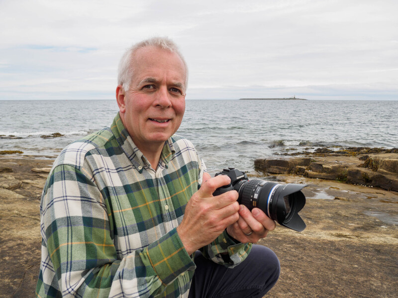 Photographer holding a camera with telephoto lens while crouching on a rocky beach.