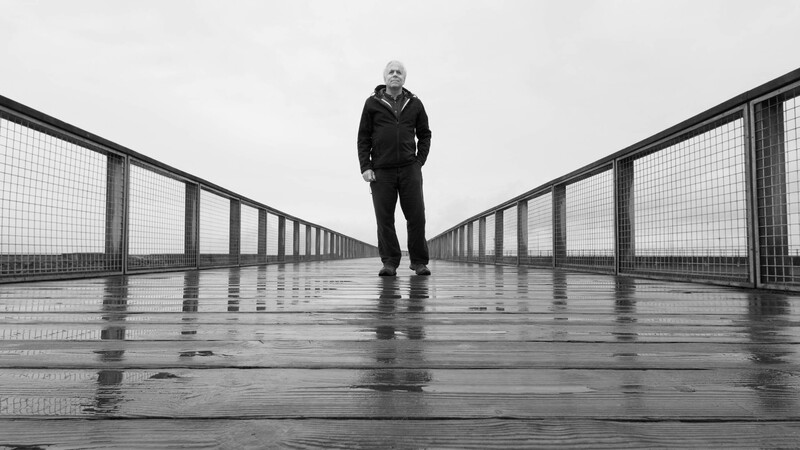 Person standing alone on a wet boardwalk with symmetrical mesh railings extending into the distance.