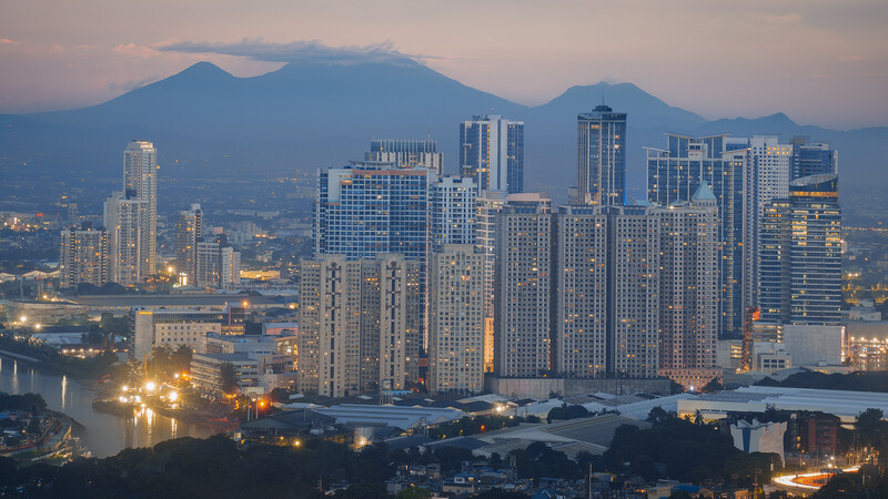 Urban skyline at dusk with illuminated high-rise buildings, a river in the foreground, and snow-capped mountains in the background.