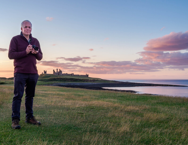 Runner jogging through coastal grassland at sunset with castle ruins visible across water in distance.