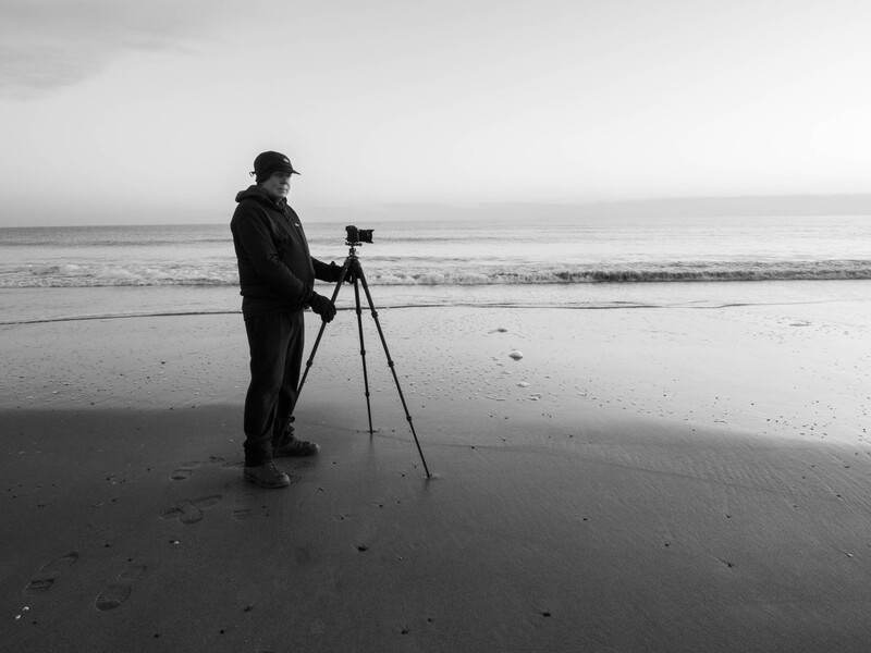 Photographer with tripod-mounted camera standing on wet beach at shoreline.
