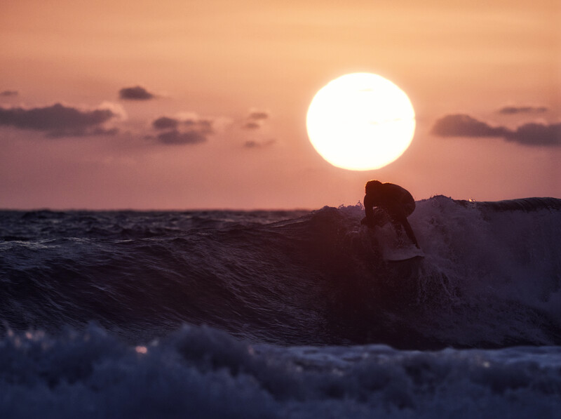 Surfer riding a wave during golden hour with a large sun on the horizon.