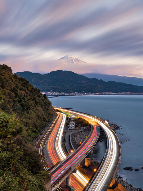 Coastal highway with light trails curving through mountainous terrain at dusk, snow-capped peak visible across the bay.