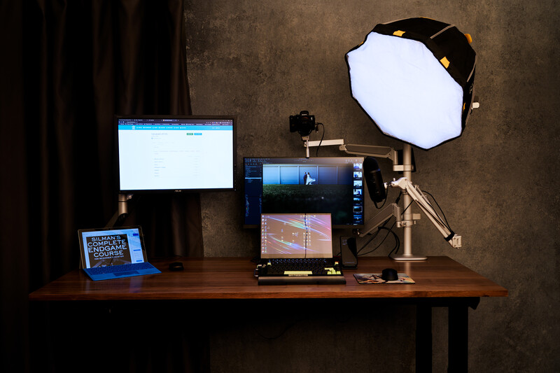 Photography workspace with monitor, laptop, camera on tripod, and softbox lighting setup on wooden desk.