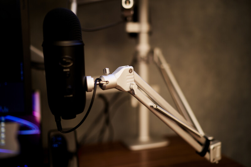 White boom arm microphone mount with articulating joints positioned next to a black condenser microphone on a desk setup.