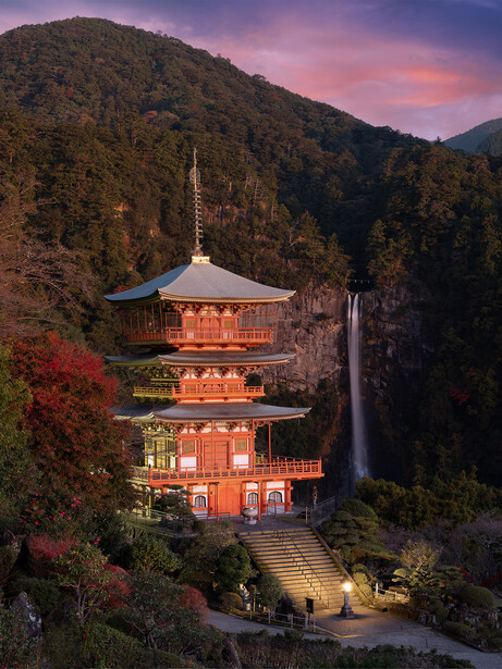 Red pagoda illuminated at dusk nestled in a forested mountain valley.