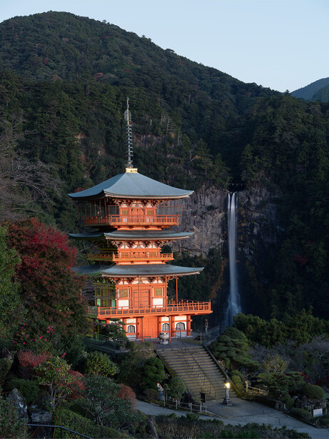 Red-orange pagoda with three tiers nestled against a forested mountainside at dusk.