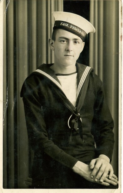 Young man in sailor uniform with cap and neckerchief, posed against curtained backdrop.