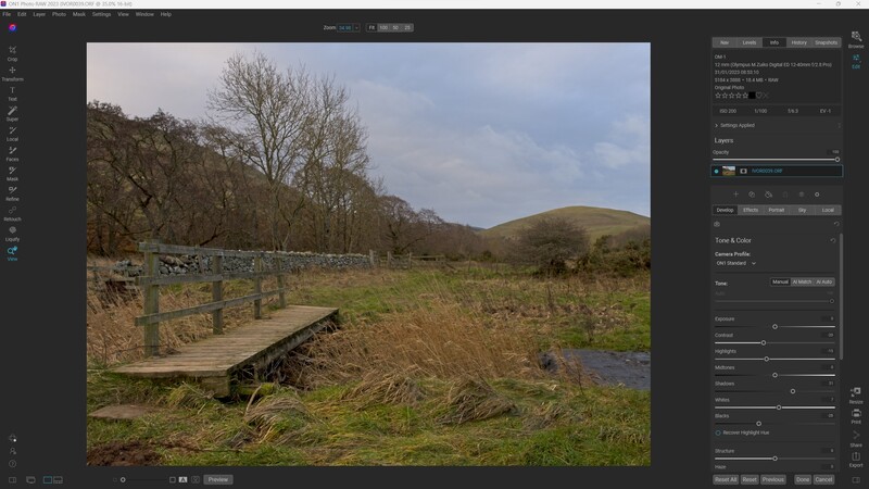 Wooden footbridge crossing a stream in a rural valley with bare trees and rolling hills.