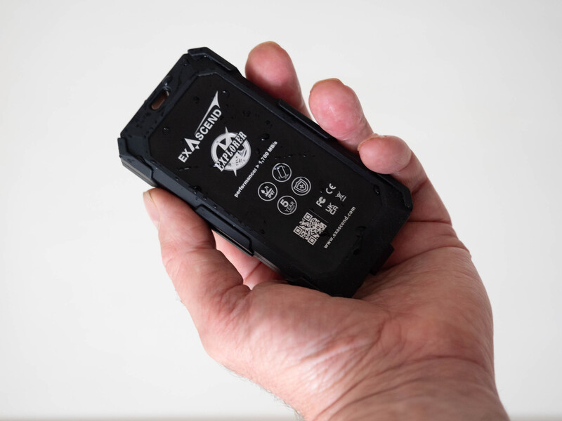 Hand holding a small black rectangular device with white text and iconography on its back surface.