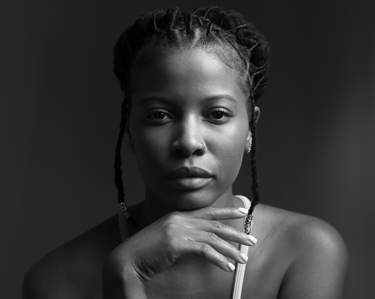 Black and white portrait of a woman with braided hair, looking downward with her hand near her chin.