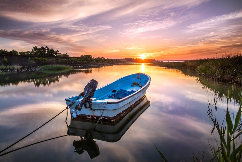 Blue boat anchored in still water at sunrise, reflected in glassy surface with reeds and trees lining the shoreline.
