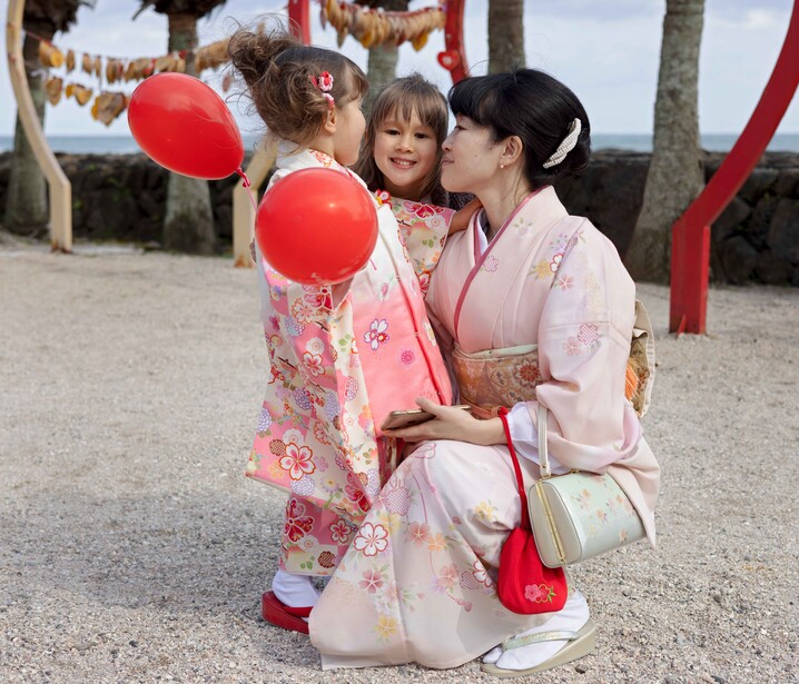 Woman in pink kimono kneeling with two young children holding red balloons at a beachside venue.