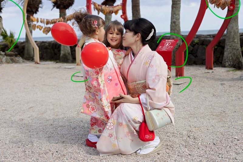 Woman in pink kimono kneeling with two young children holding red balloons at a beach celebration.