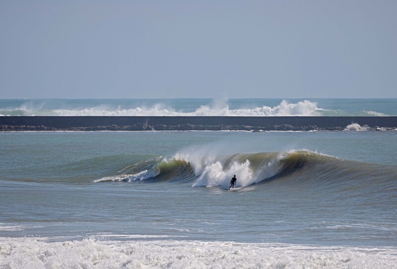 Surfer riding a large wave with white foam crest in bright daylight, other wave lines visible beyond.