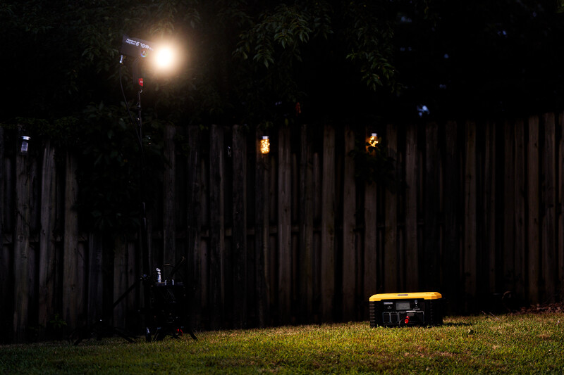 Nighttime scene with a bright work light illuminating a wooden fence and grassy area with a yellow utility vehicle.
