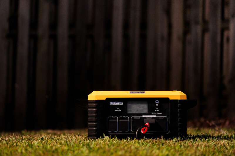Portable power station with yellow and black casing positioned on grass with dark wooden fence background.