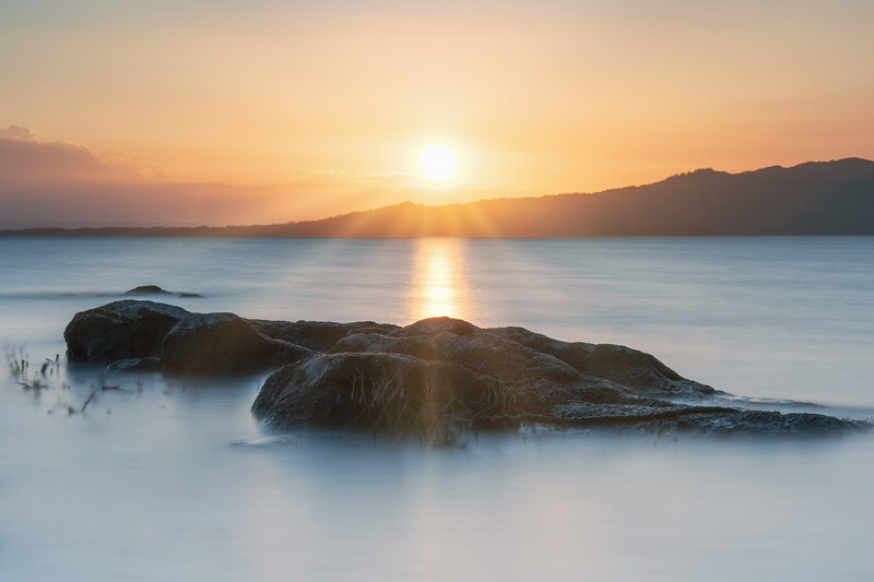 Silhouetted rocks in a misty sea with a golden sunrise reflecting across calm water and mountains in the distance.