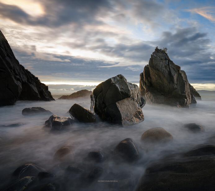 Rocky sea stacks rising from misty ocean water at twilight with dramatic clouds above.