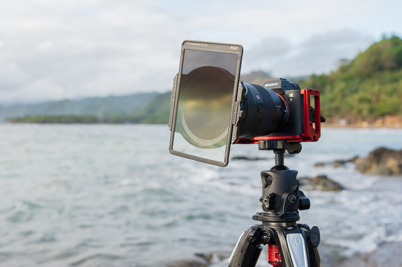 A smartphone mounted on a tripod with a reflector panel in front of a red camera, positioned overlooking a calm lake with forested hills in the background.