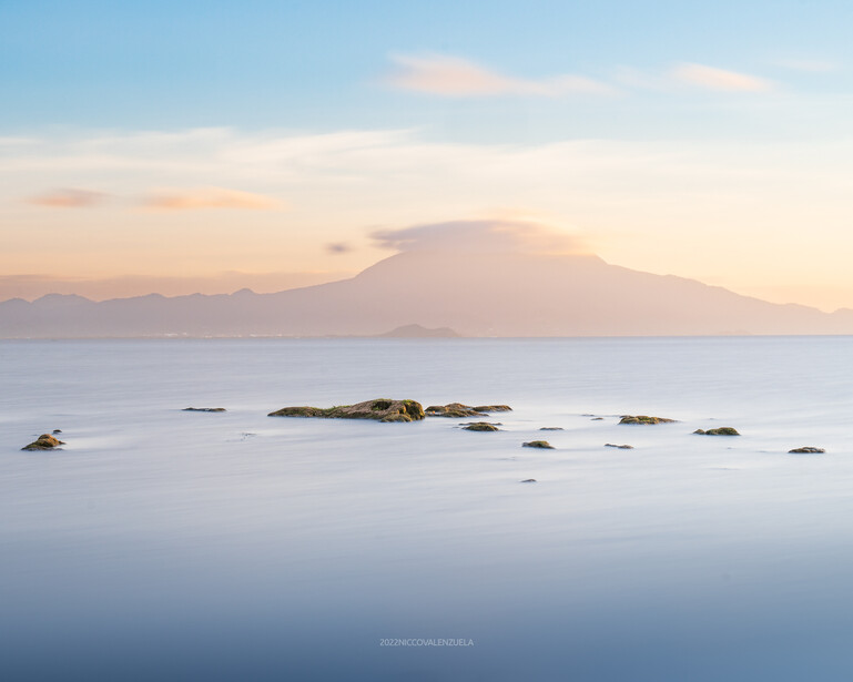 Mountain peak reflected in still water at sunrise with scattered rocks in foreground.
