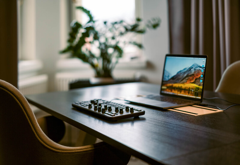 Music production setup on a desk with a MIDI controller, laptop displaying mountain landscape, and potted plant in natural window light.