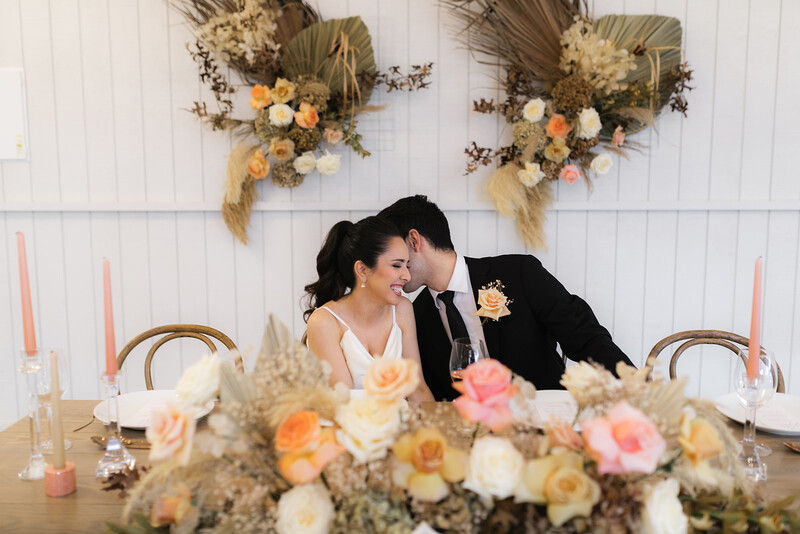 Groom kissing bride's cheek at reception table with floral arrangement and dried flower wall installation.