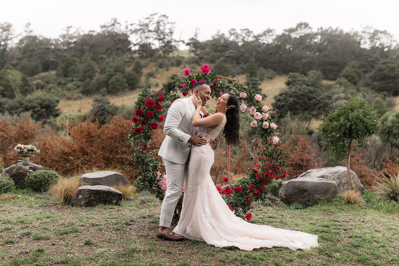 Bride and groom kissing under a floral arch in an outdoor garden setting.