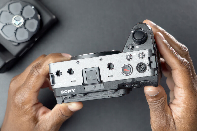 Hands holding a Sony compact camera with white body and black accents, viewed from above.