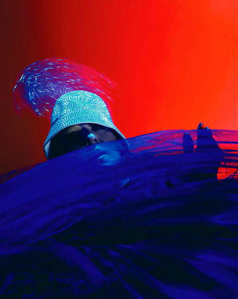 Person wearing textured turquoise garment on blue-toned rocks against red sky.