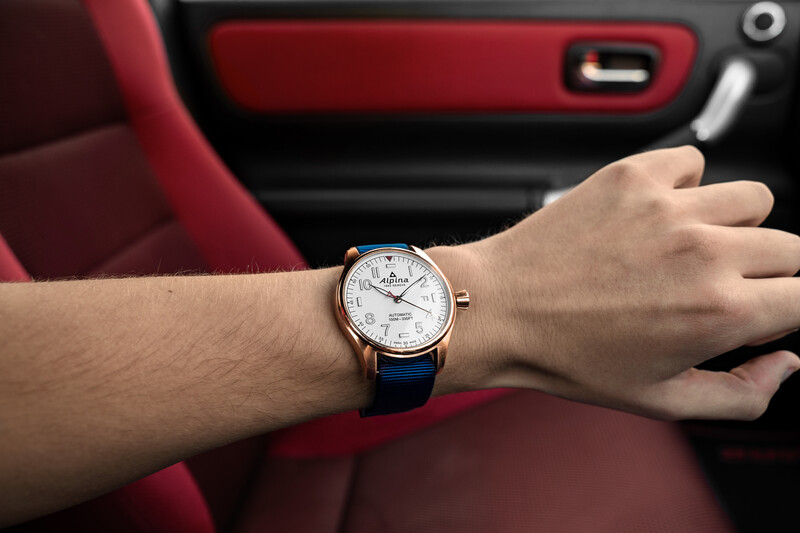 Man's wrist wearing a silver-cased watch with dark leather strap inside a red car interior.