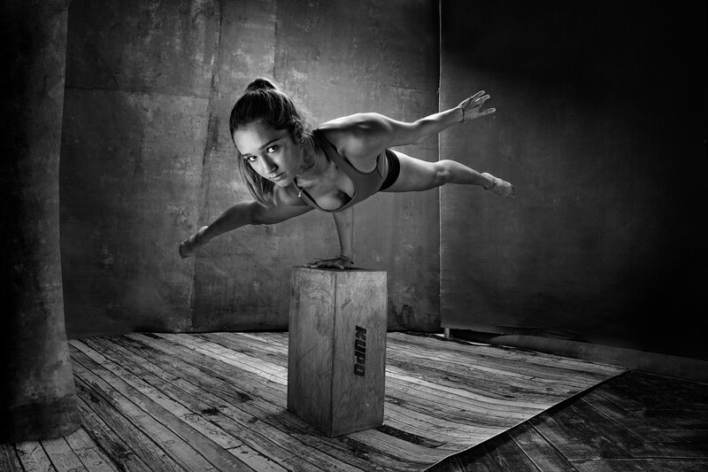 Shirtless male athlete performing a one-armed handstand on a wooden box in a minimalist industrial studio.