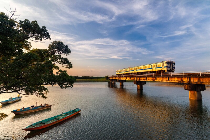 Yellow and orange train crossing a bridge over a calm river with moored boats in the foreground.