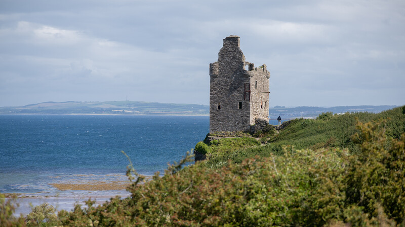 Ruined stone tower on a grassy coastal headland overlooking blue water and distant hills.