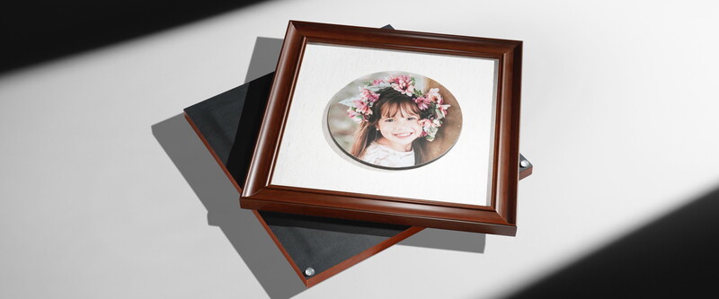 Framed oval portrait of a smiling child wearing a floral crown, displayed on a white surface with natural sunlight casting shadows.