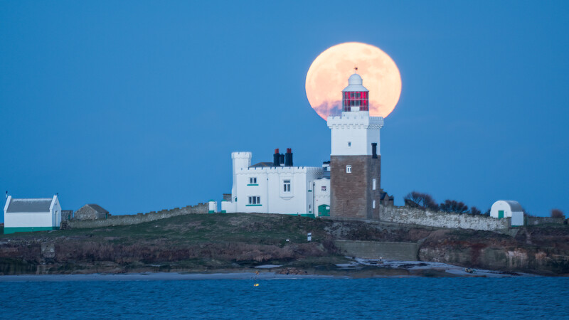 Coastal lighthouse on rocky headland silhouetted against a full moon at dusk.