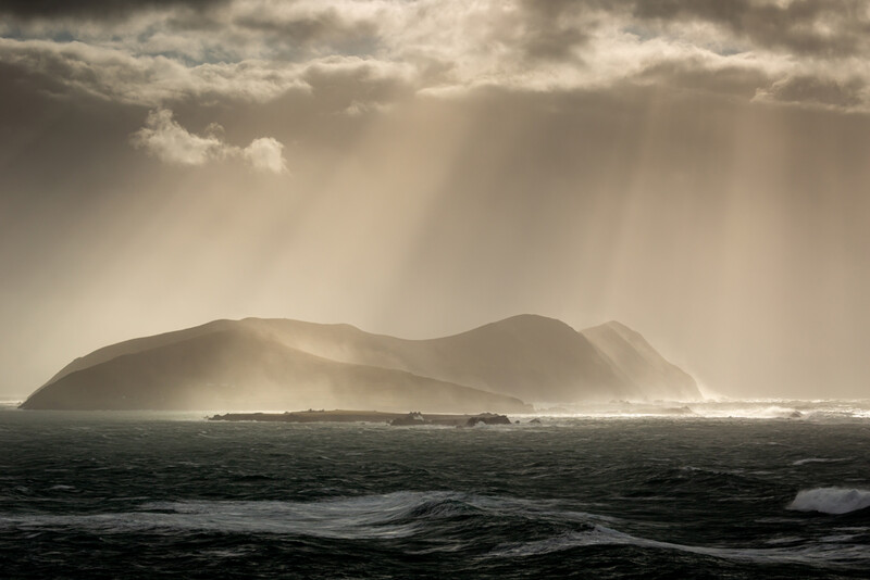Volcanic island shrouded in mist under dramatic cloudy sky with crepuscular rays.