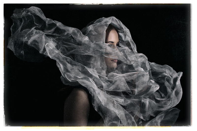 Person's face partially obscured by flowing, translucent fabric against a dark background.