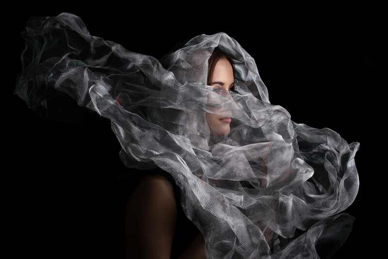 Person's face partially obscured by flowing, translucent gray tulle fabric against a black background.