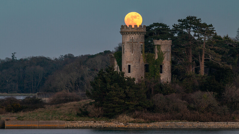 Historic stone tower ruins silhouetted against a full moon rising over a river at dusk.