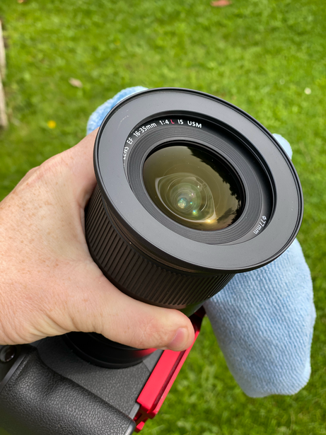 Hand holding a wide-angle camera lens with focus ring and depth-of-field markings visible against grass background.