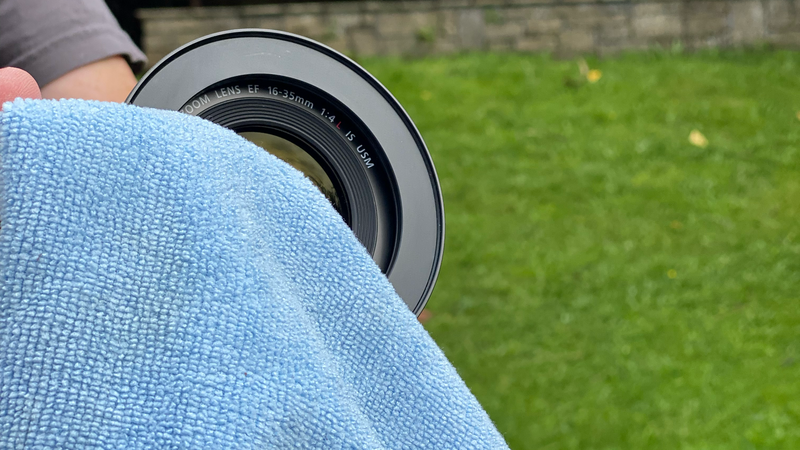 A camera lens being cleaned with a light blue microfiber cloth outdoors.